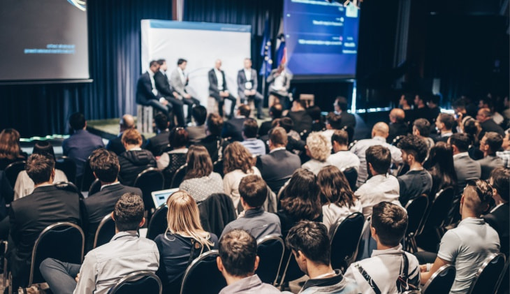 Adults at a business conference watch a group of speakers on stage